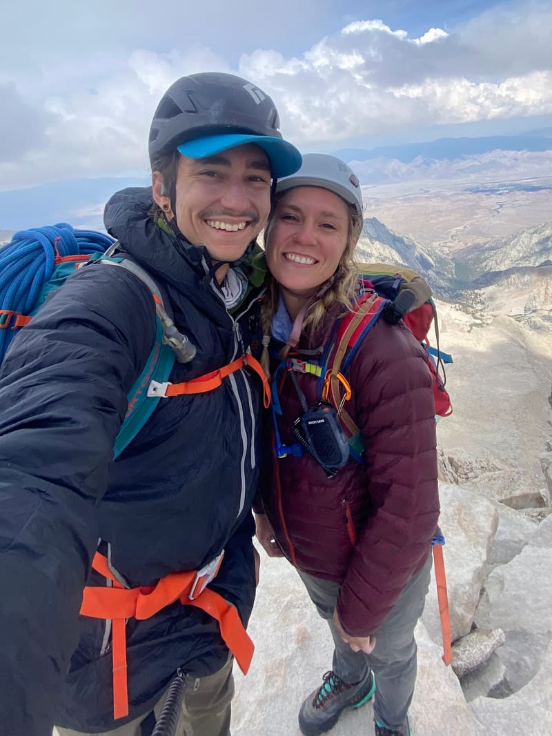 Riley and Anthony, your hosts, smiling in climbing helmets on a rocky summit with mountains and valley behind them